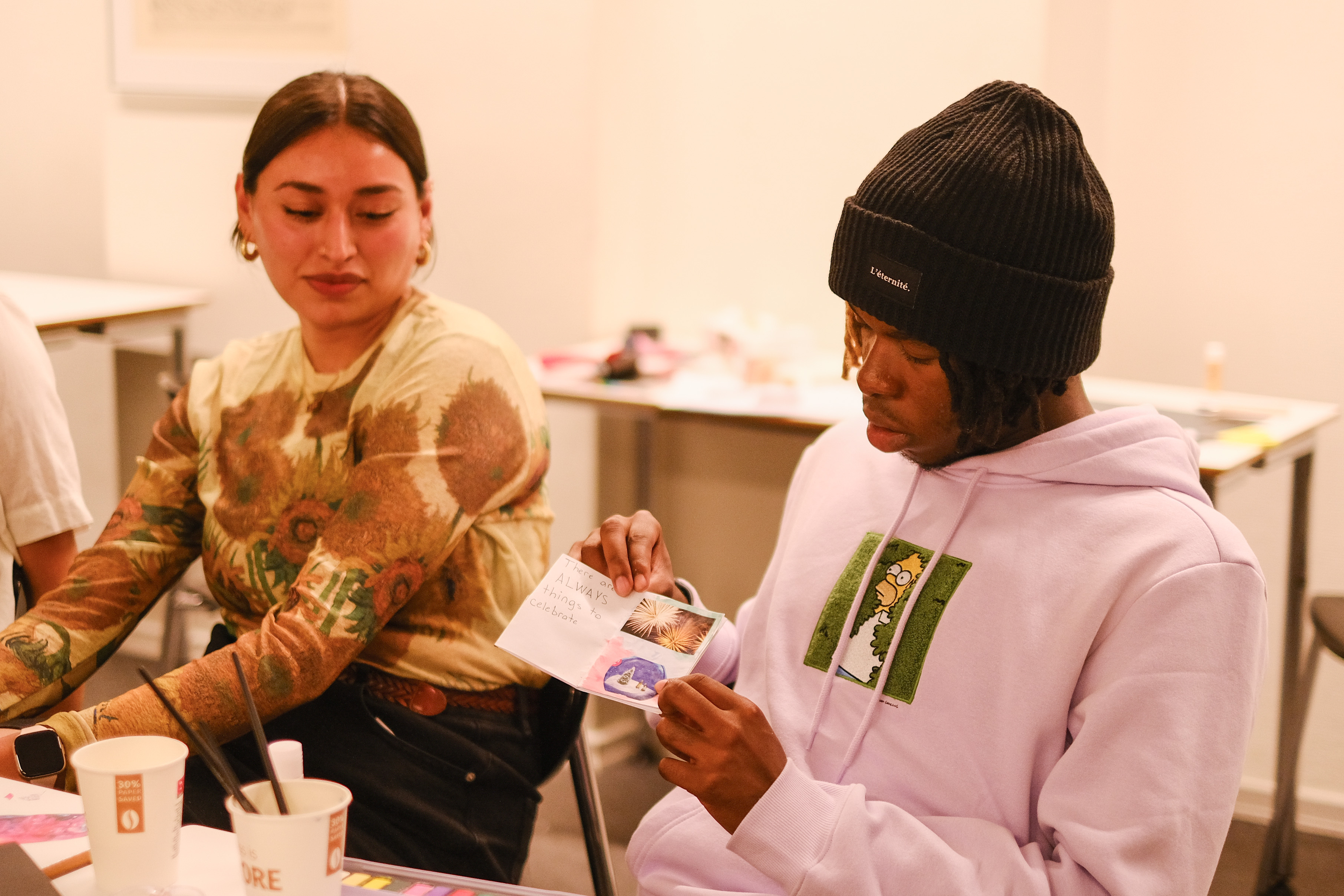 Two students sit together, while one presents his zine to her peers.